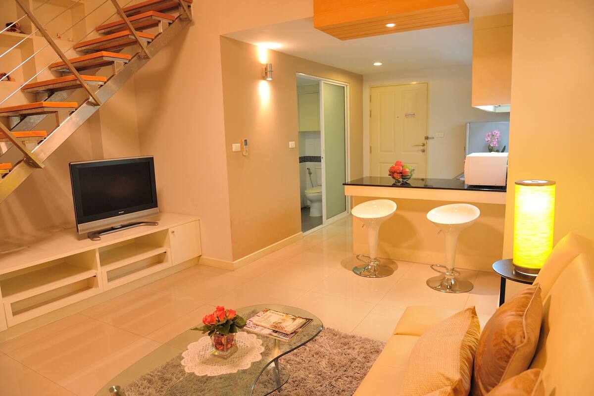 A modern living room with a TV, a glass coffee table with a vase, a sofa, and a staircase.