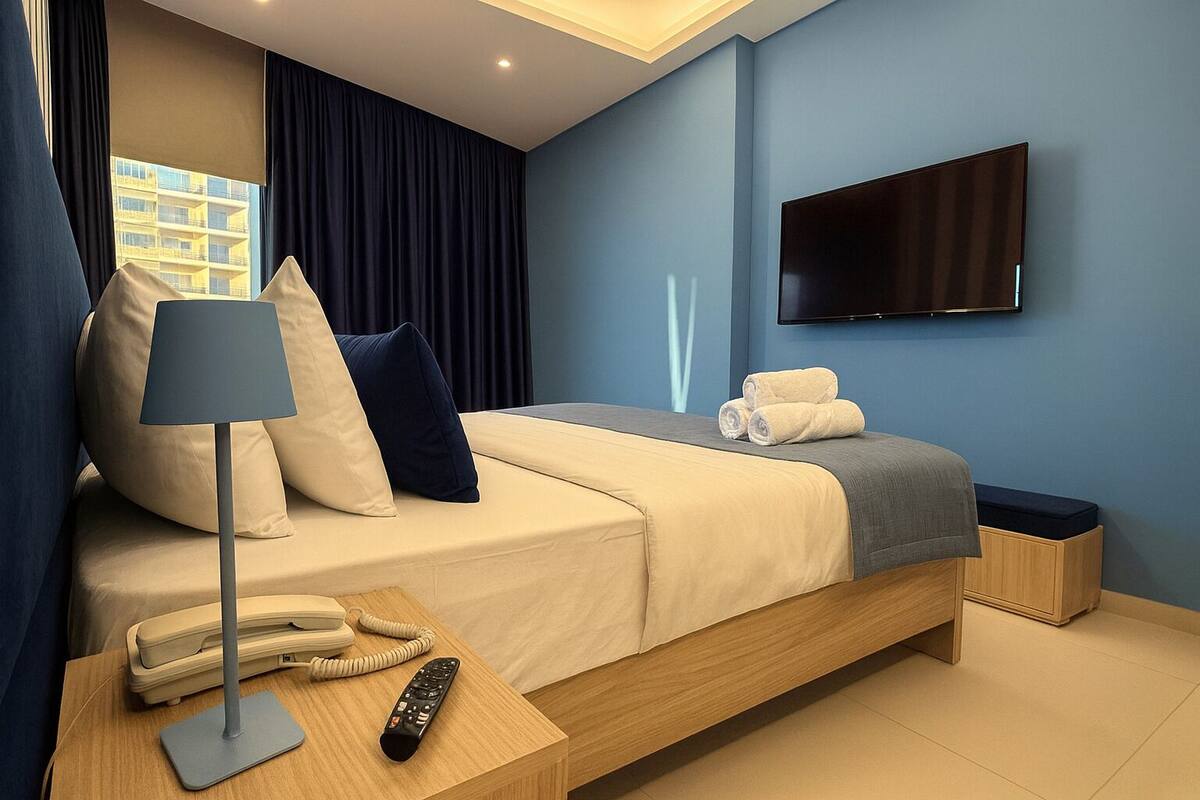 A modern hotel room with a bed, a TV, a bedside table with a lamp, a phone, and a remote control.