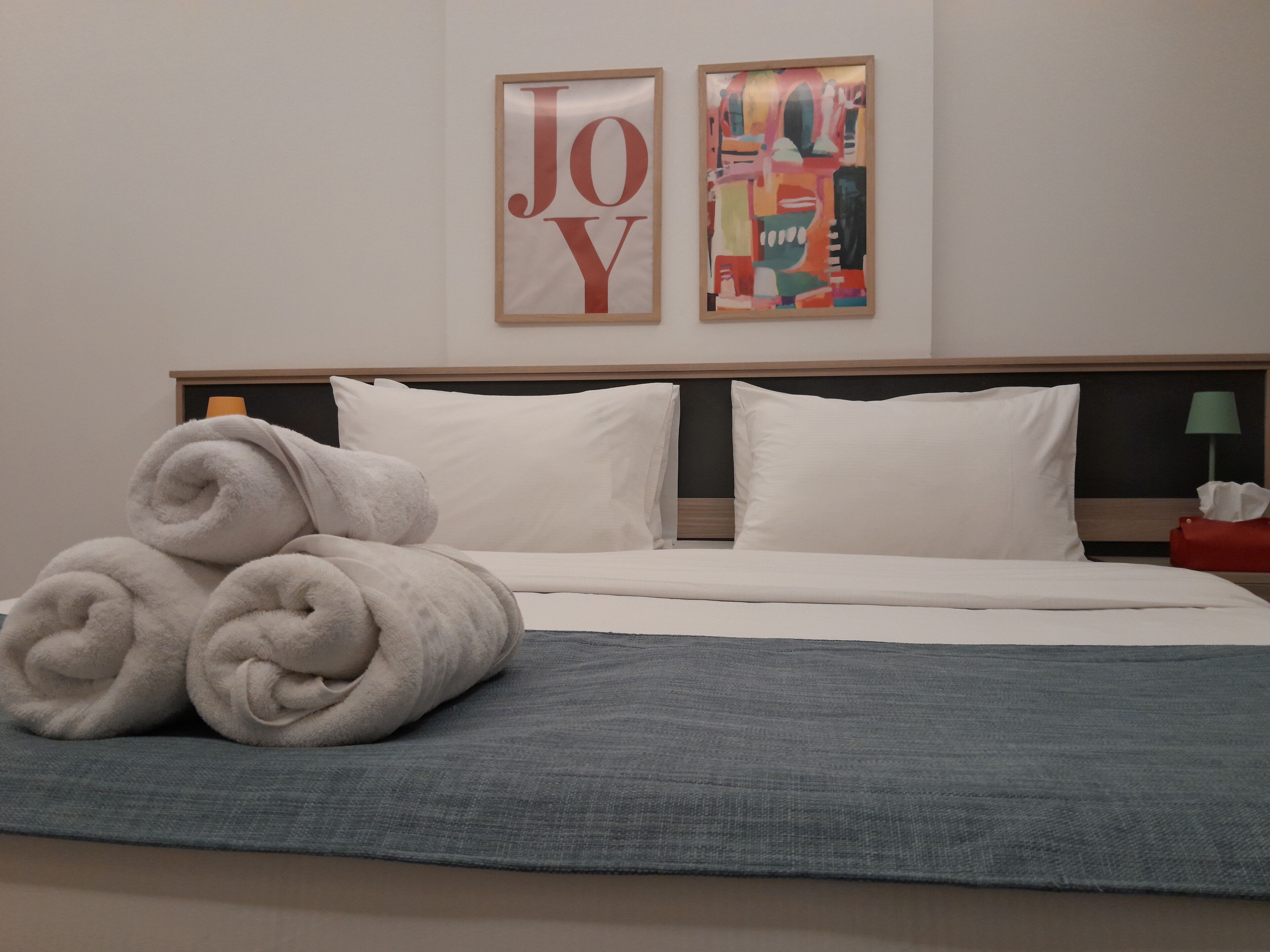 A neatly made bed with white linens, a grey blanket, and rolled towels. There are two pillows and a headboard with a wooden frame. Two framed artworks are hung on the wall above the bed.