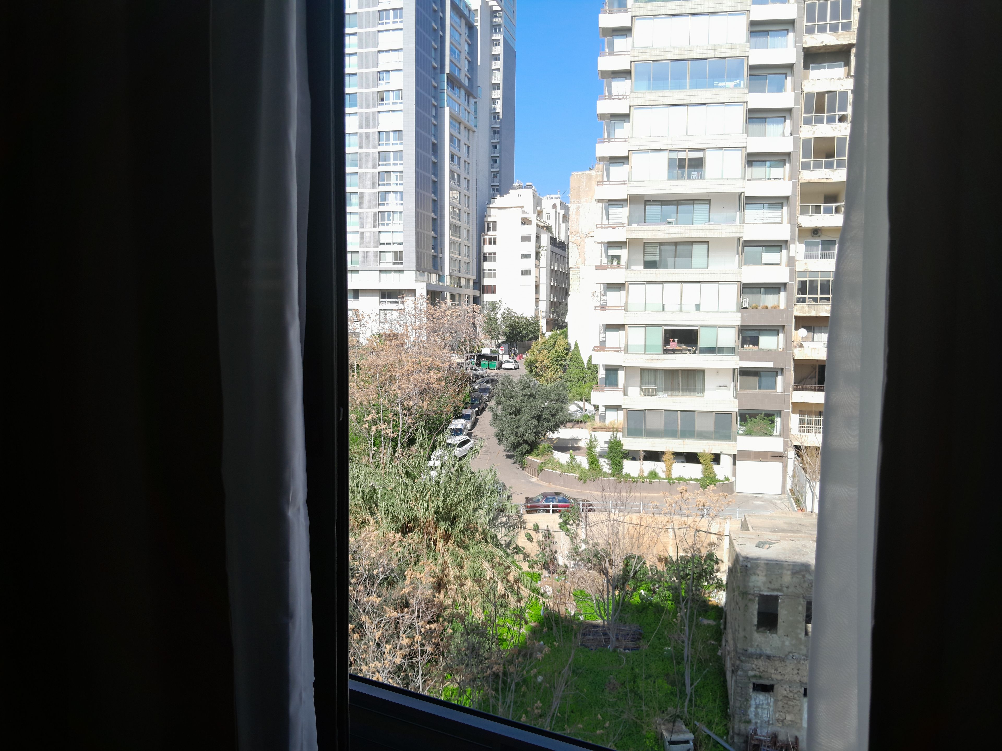 Vista de una zona residencial con edificios altos, una calle con coches estacionados y algo de vegetación.