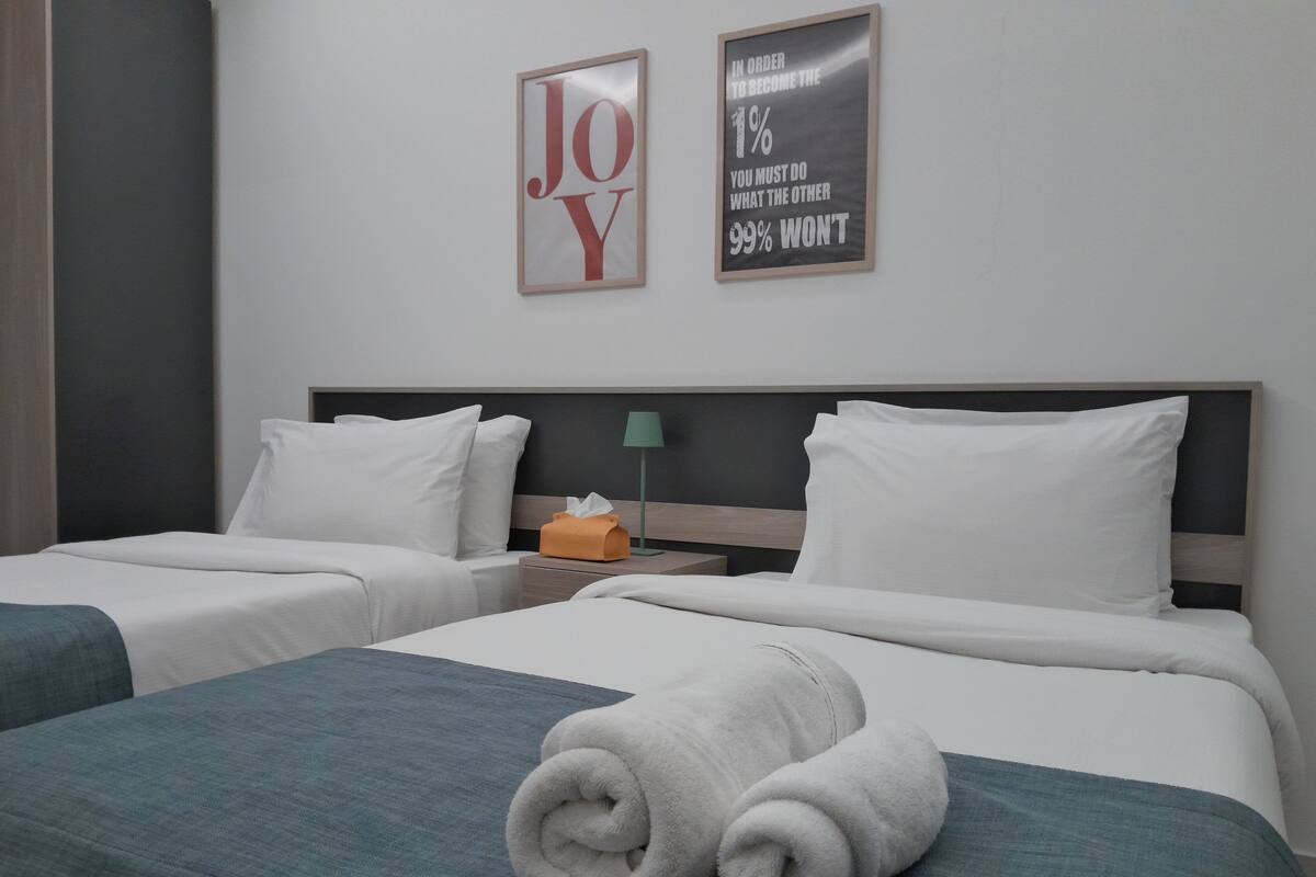 A hotel room with two beds, each with white bedding and teal throw pillows. There are towels on the beds and a framed motivational quote on the wall.