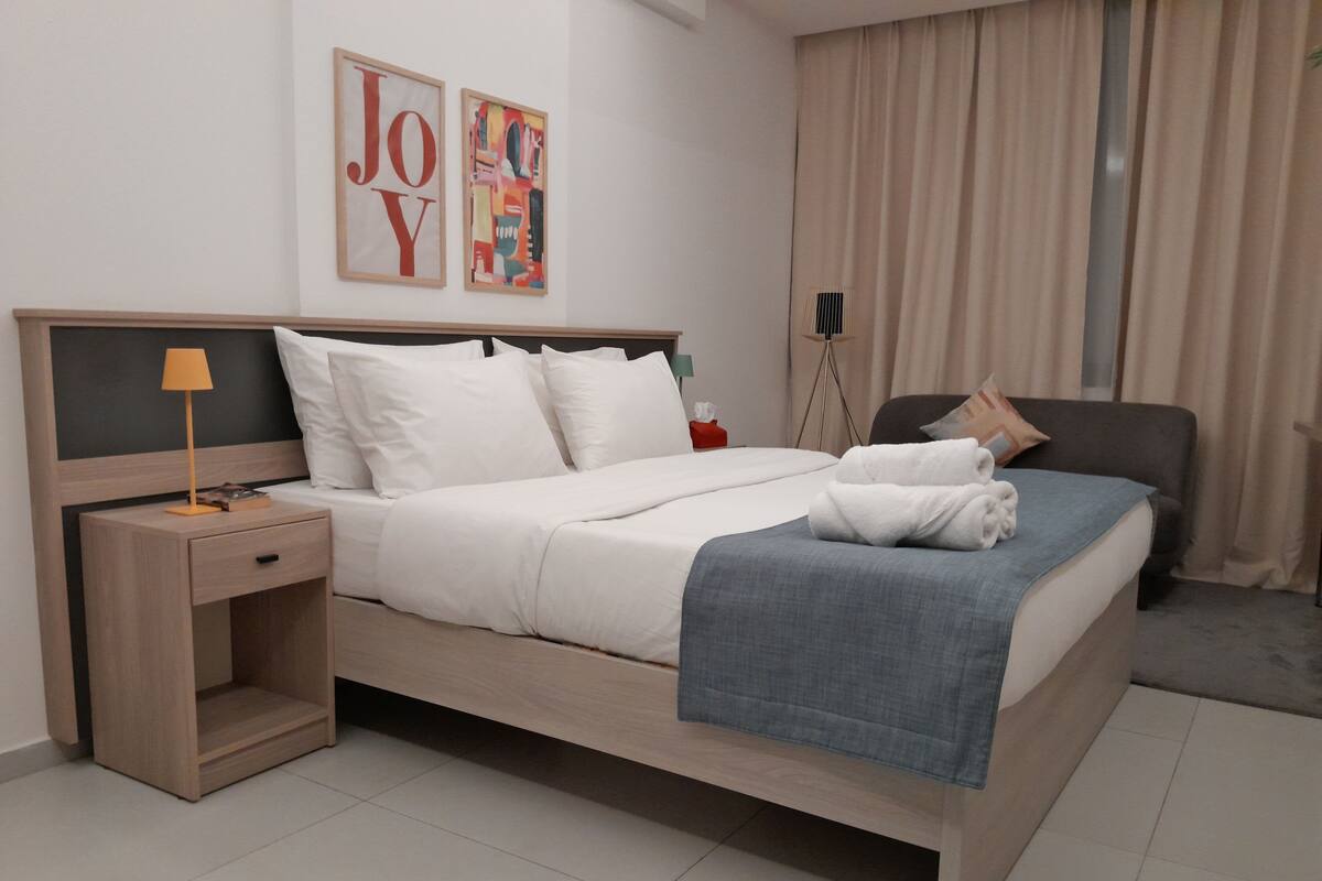 A modern hotel room with a bed, a nightstand, a lamp, a chair, and a window with curtains.