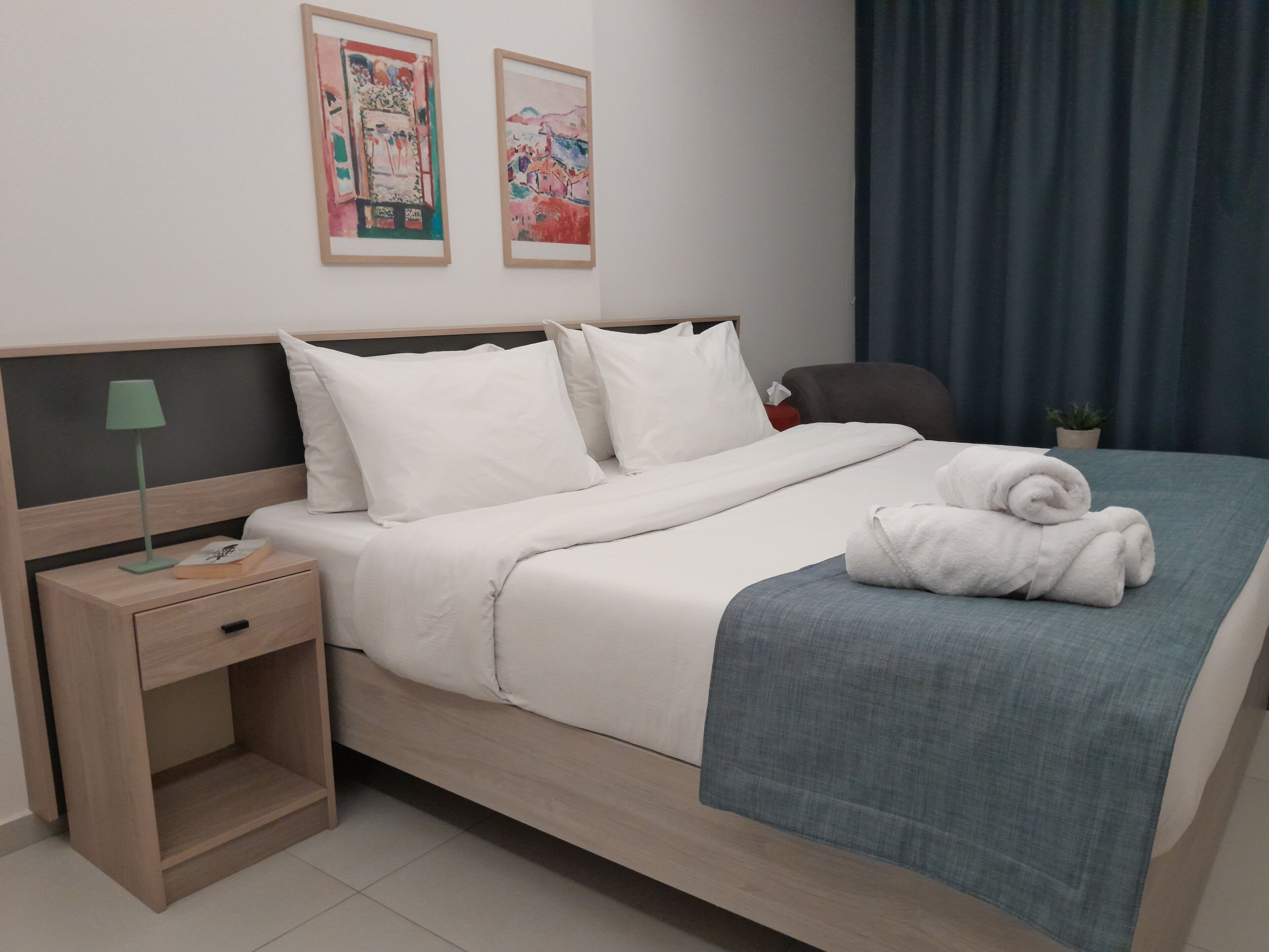 Una cama bien hecha con sábanas blancas, una lámpara de noche, una mesita de noche pequeña y una planta en maceta.