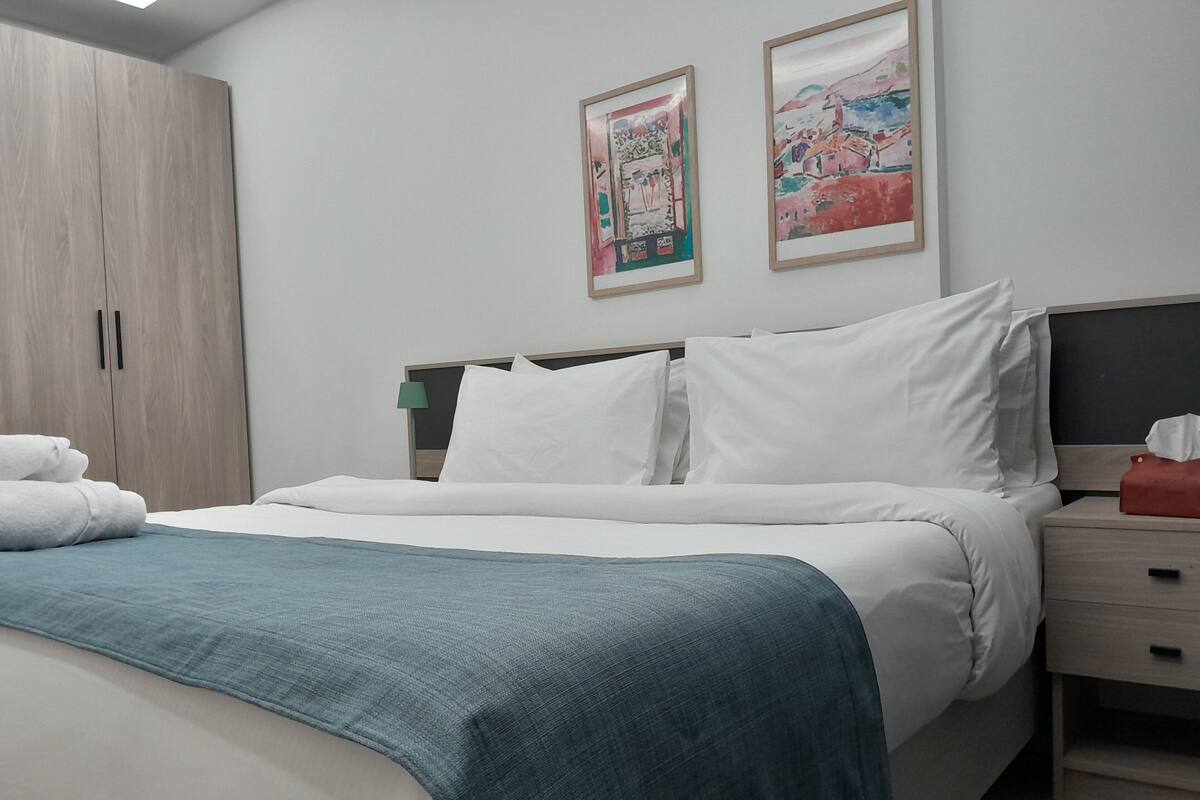 A neatly made bed with white linens and a teal throw blanket, a wooden wardrobe, and two framed artworks on the wall.