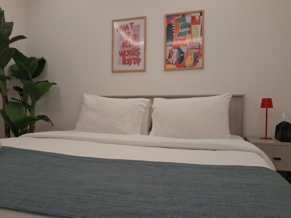 Una cama bien tendida con sábanas blancas, un plaid gris y una planta verde grande a su lado. Dos cuadros están colgados en la pared sobre la cama.