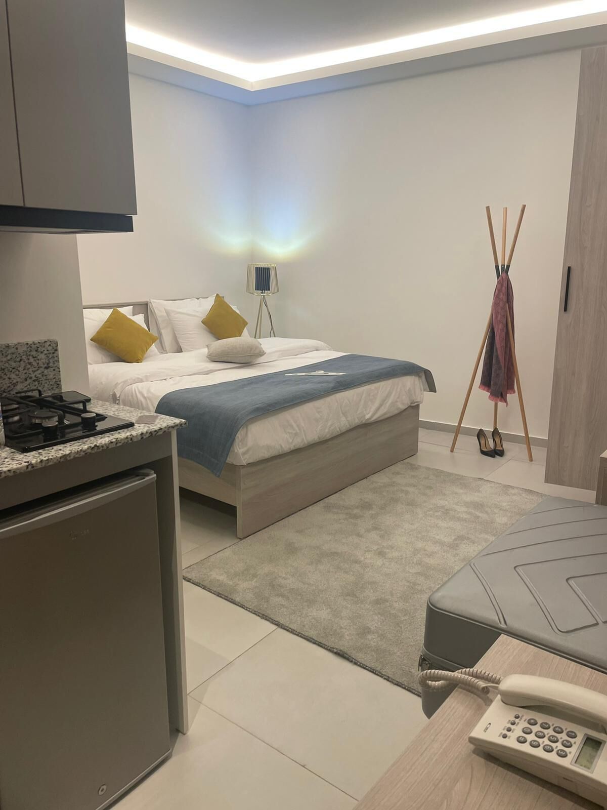 A modern hotel room with a bed, bedside table, a small kitchenette, a bedside lamp, and a telephone.