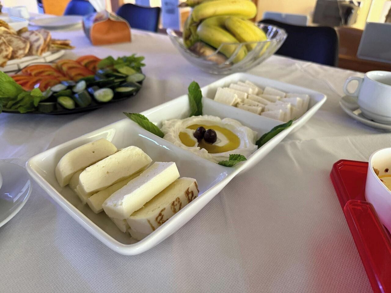 Et bord med et udvalg af mad, blandt andet skivede bananer, en skål med oliven og en gul sauce, samt en tallerken med skivede agurker og tomater.