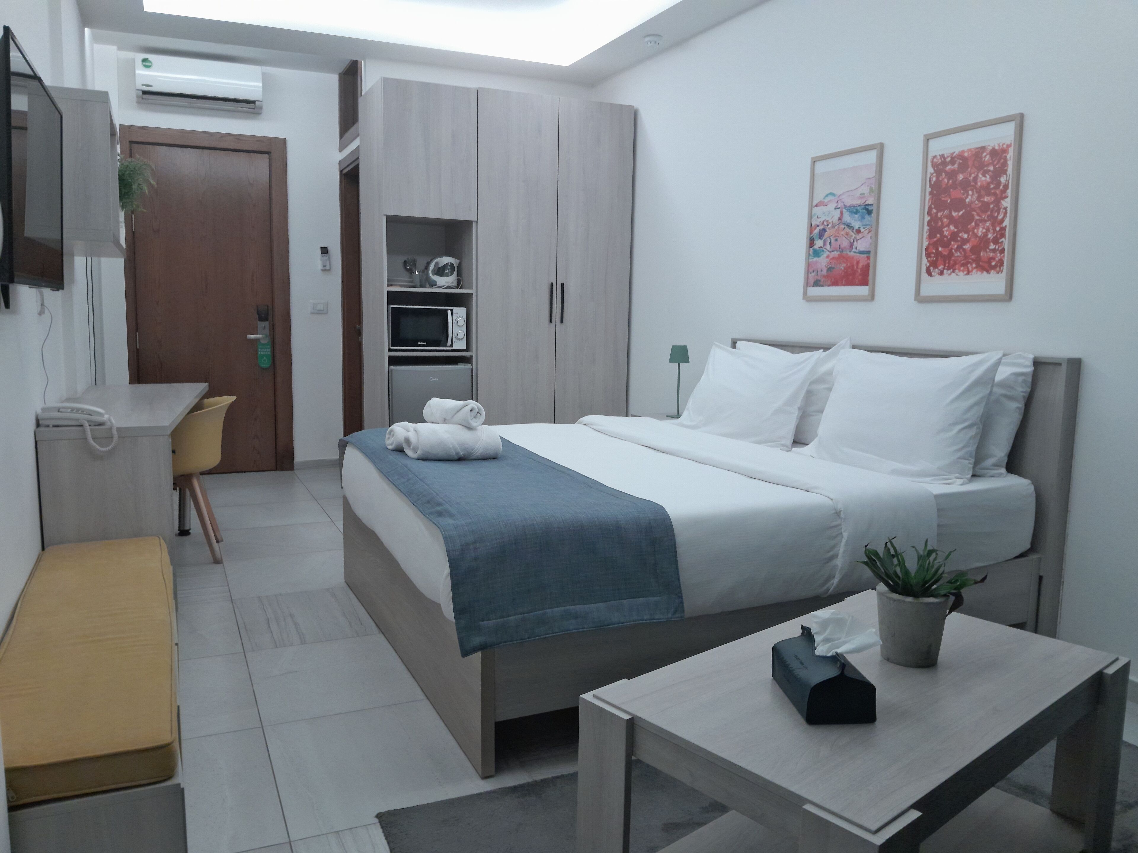 A modern hotel room with a bed, a desk, a chair, a small table, and a potted plant.