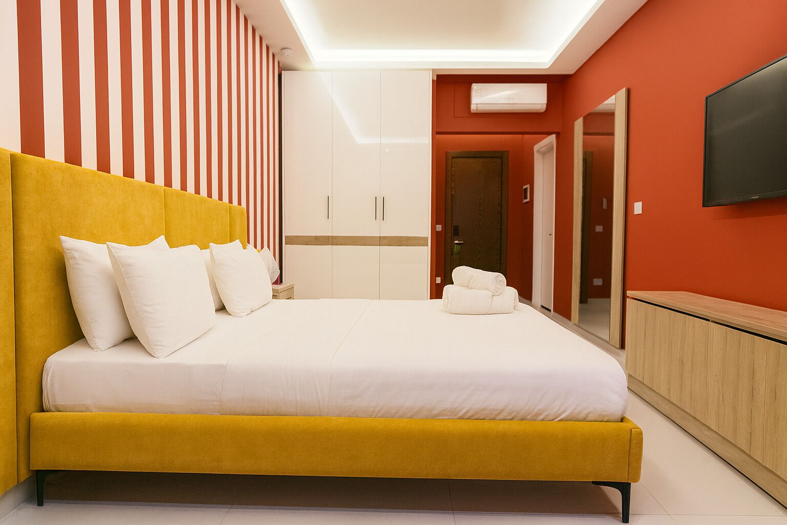 A modern hotel room with a yellow bed, white pillows, a striped wall, a wooden cabinet, and a TV on the wall.