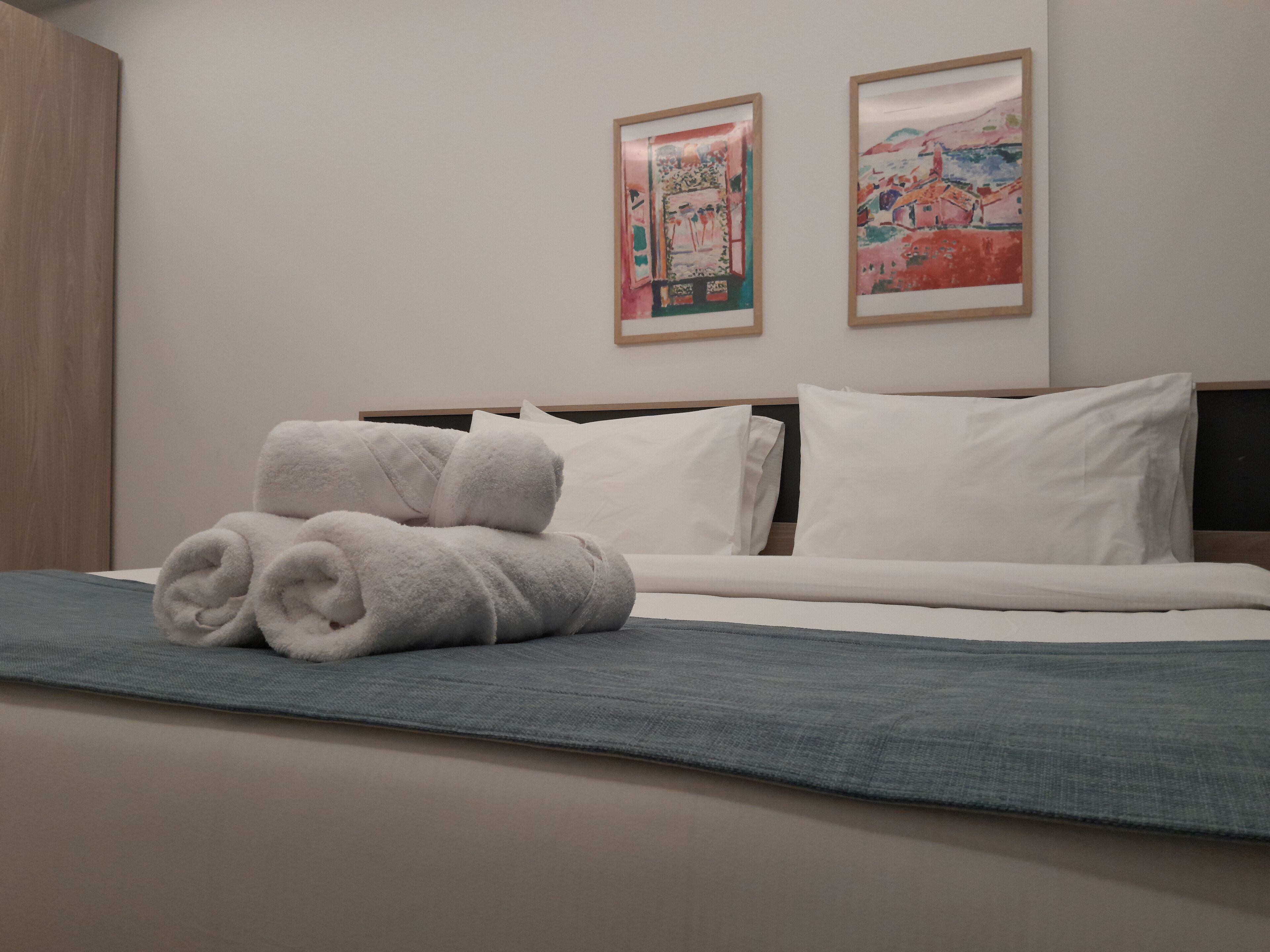 A neatly made bed with white pillows and a blue blanket, two framed artworks on the wall, and a wooden wardrobe to the left.