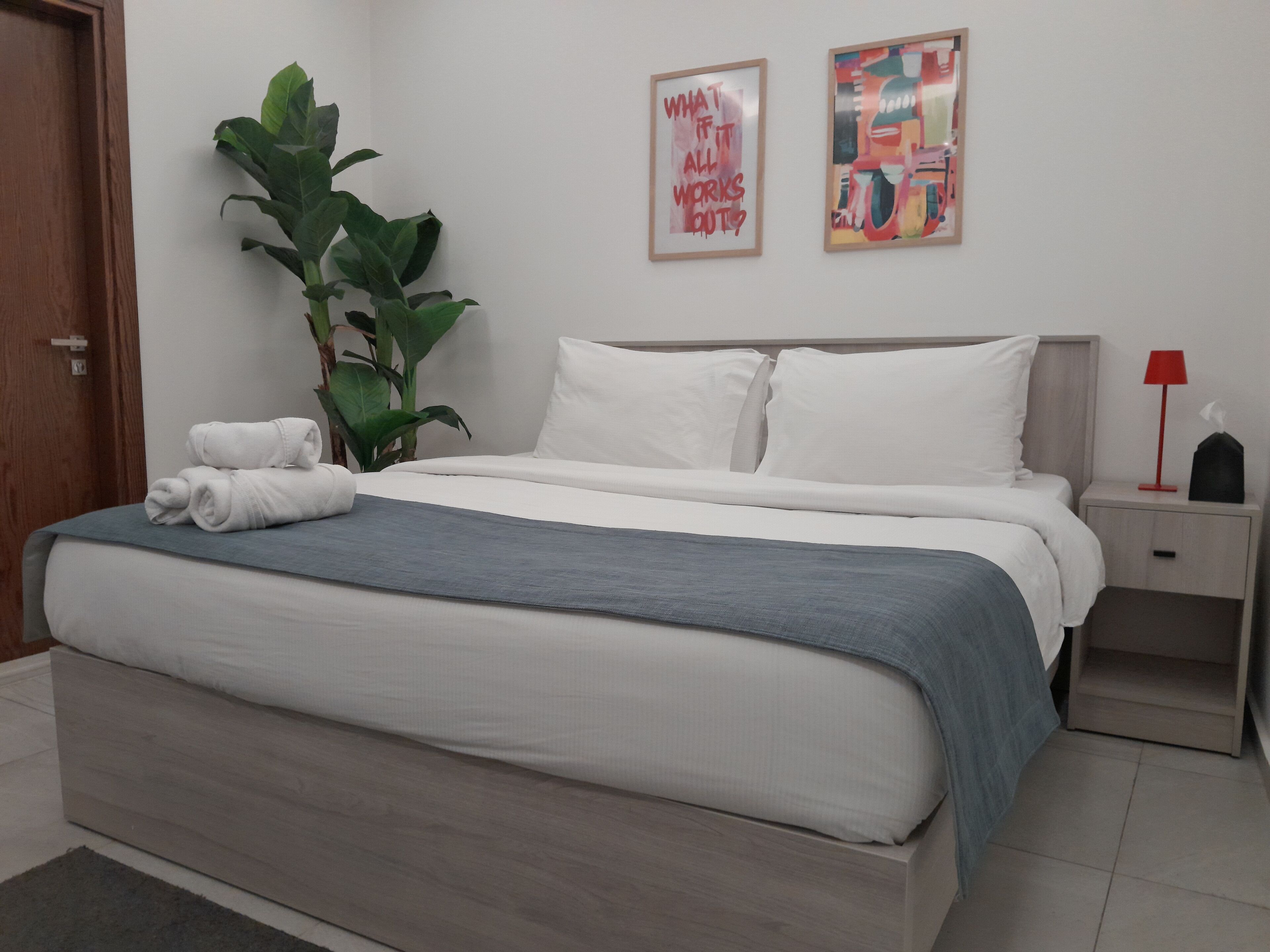 A neatly made bed with white and gray bedding, a large green plant beside it, and two framed artworks on the wall.