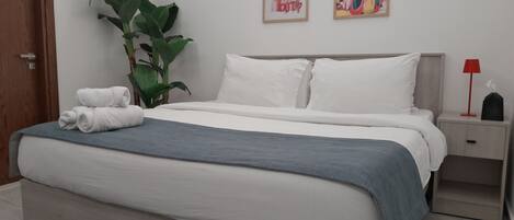 A neatly made bed with white and gray bedding, a large green plant beside it, and two framed artworks on the wall.