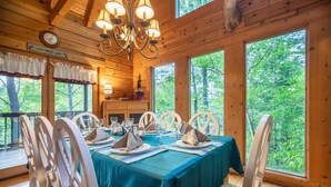 Une cabane en bois avec une table à manger dressée pour quatre personnes, entourée de chaises blanches, de grandes fenêtres donnant sur la forêt et un lustre.