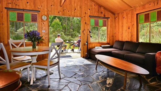 Een houten cabin met een stenen vloer, een eettafel met stoelen, een bank en uitzicht op het bos door grote ramen.