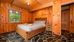 Een houten kamer met een stenen vloer, een bed met houten hoofdeinde, een raam met luxaflex en een deur.