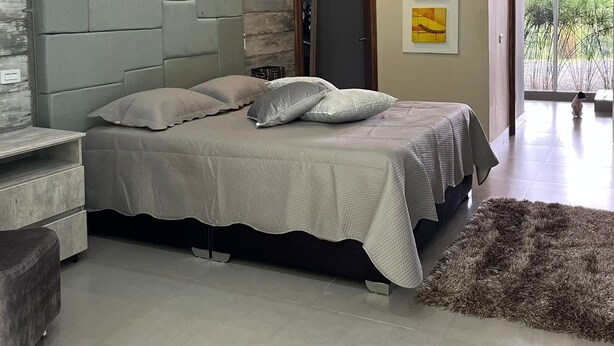 Une chambre à coucher moderne avec un lit, un banc recouvert d’un tissu gris, un tapis gris et un climatiseur fixé au mur.