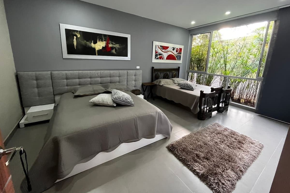 Ein modernes Schlafzimmer mit einem großen Bett, einem kleineren Bett, einem Balkon mit Blick auf Grünflächen und zwei Kunstwerken an der Wand.