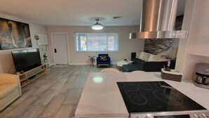 Una cocina moderna con placa de inducción, horno integrado y cafetera. Hay un comedor con mesa y sillas, y una sala de estar con sofá y televisor.