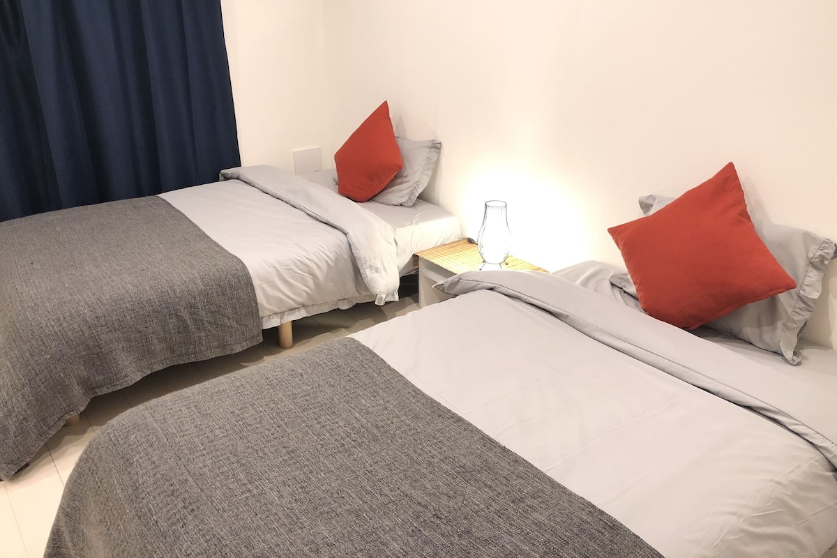 A room with two beds, each with grey and red bedding, a bedside table with a lamp, and a curtain on the left.