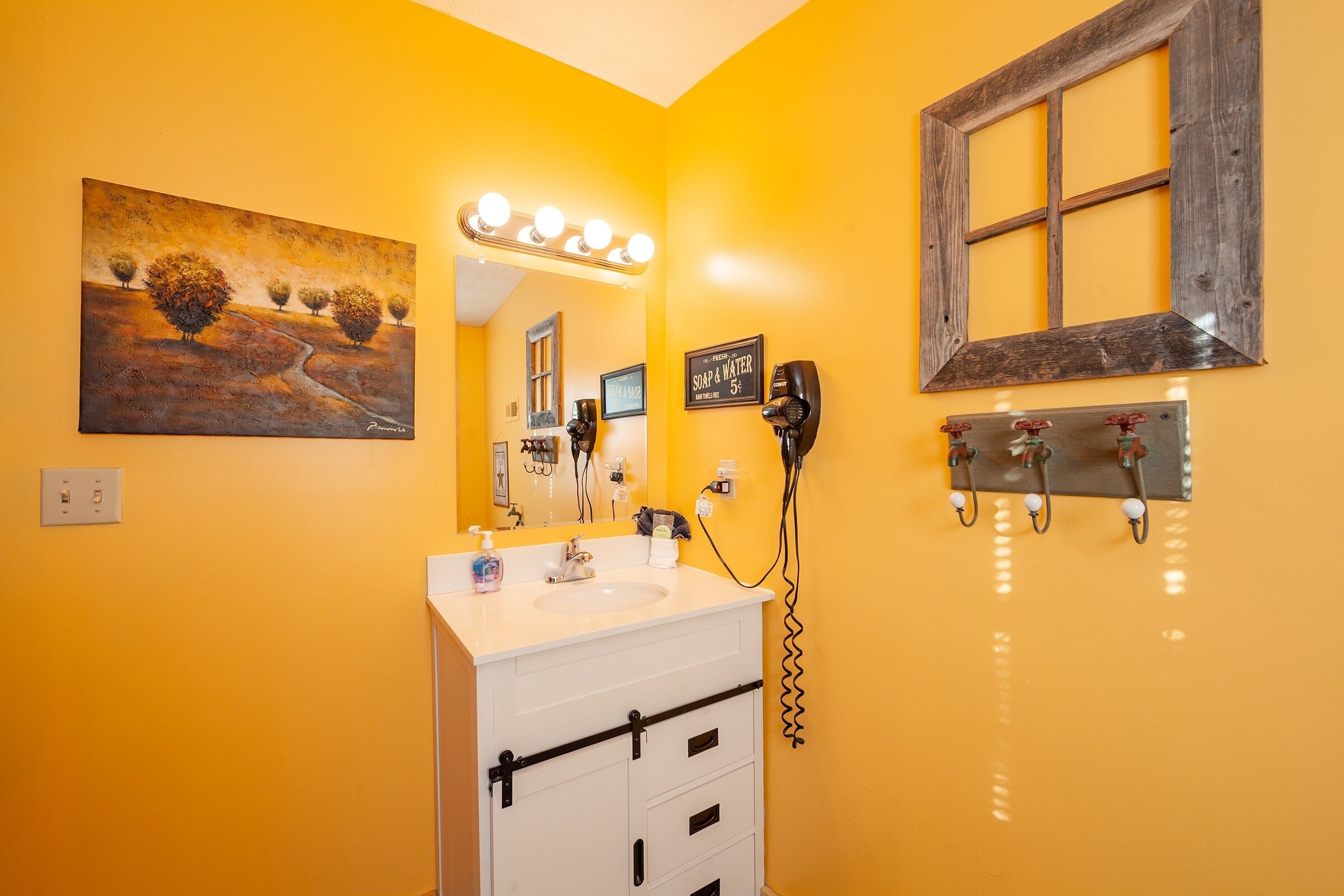 A bathroom with a yellow wall, a white vanity, a mirror with lights, a hairdryer, and a painting.