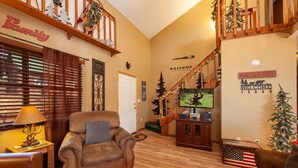 A cozy living room with a leather armchair, a wooden coffee table, and a television on a wooden stand. A staircase with a railing and a Christmas tree is visible in the background.