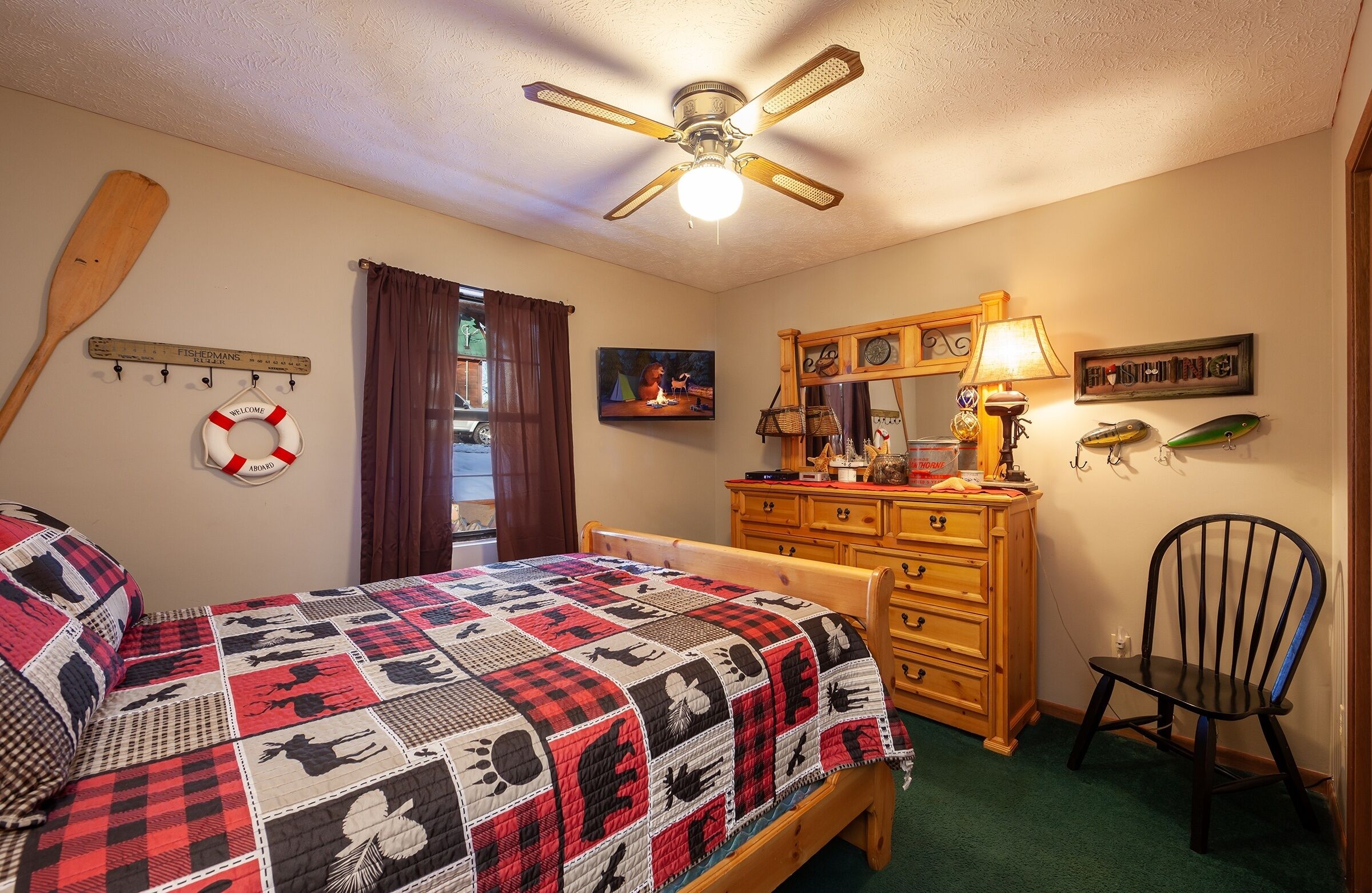 A bedroom with a bed, wooden dresser, chair, ceiling fan, and various wall decorations including a life preserver, paddle, and fish.