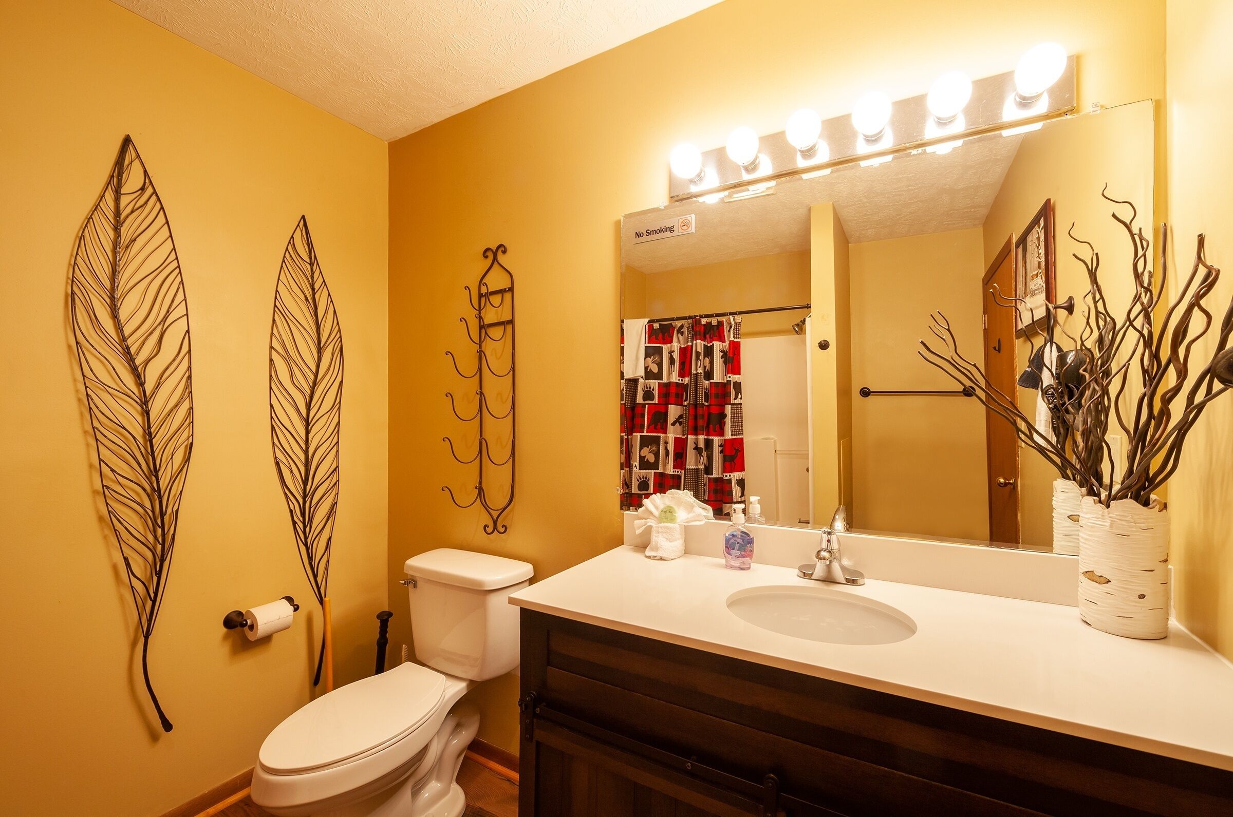 Un baño con un lavamanos blanco, un espejo con iluminación, un inodoro y decoración de hojas en la pared.