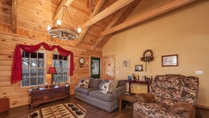 A cozy log cabin interior with a wooden ceiling, a sofa, a patterned armchair, and a small table with a lamp. - Sleepin' Inn 2 Bedroom Cabin by RedAwning (Sevierville)