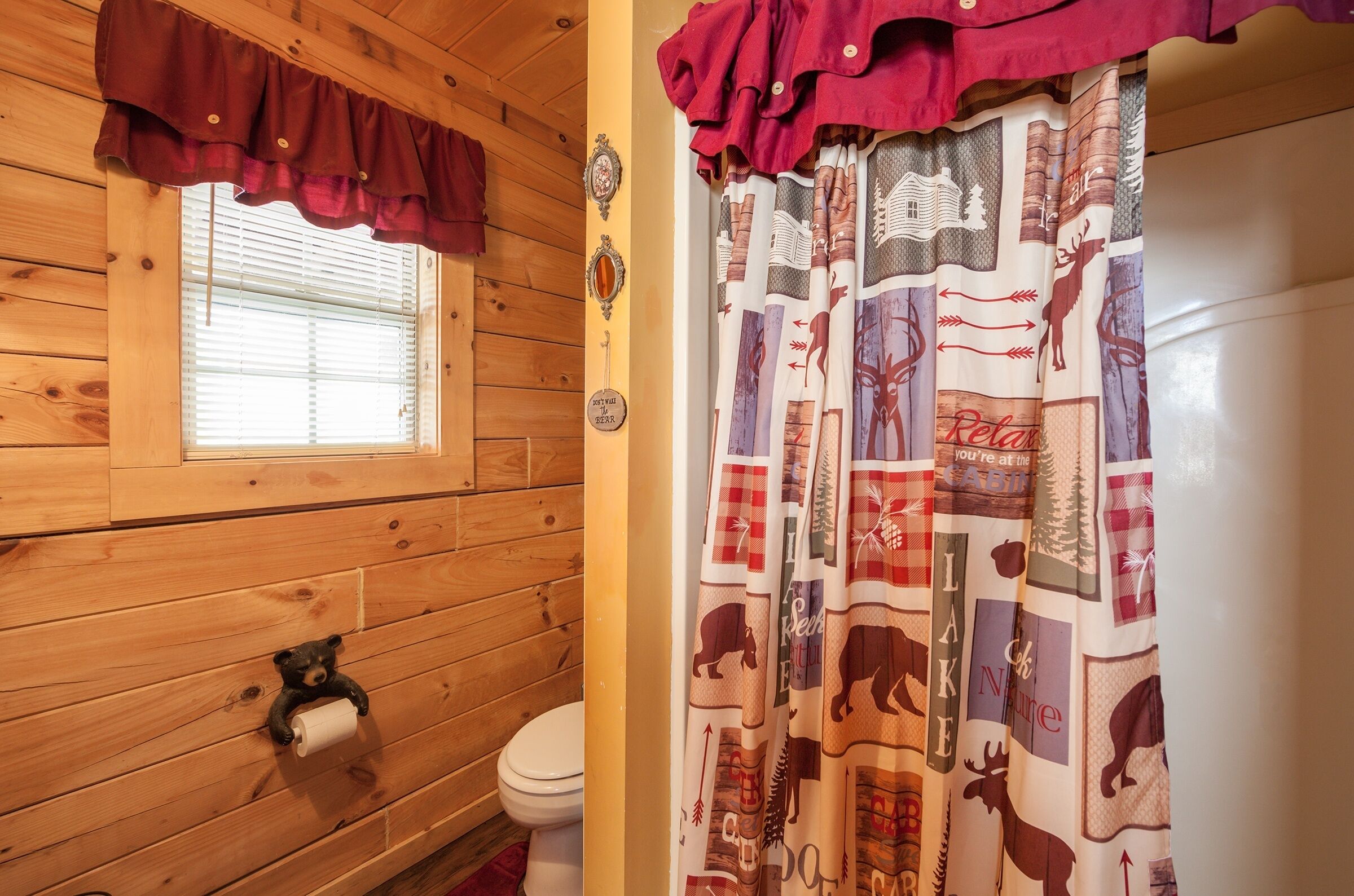 Une salle de bain rustique, aménagée dans une cabane en bois, comprenant des toilettes, une fenêtre avec stores, un porte-rouleau de papier toilette en forme d’ours et un rideau orné d’un motif inspiré de la nature.