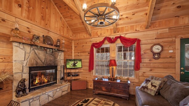 A cozy log cabin interior with a stone fireplace, wooden walls, a wooden ceiling with a wheel light, a television, and a sofa with a patterned rug. - Sleepin' Inn 2 Bedroom Cabin by RedAwning (Sevierville)