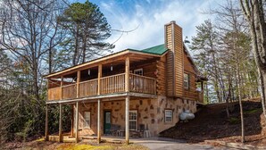 A log cabin with a balcony, stone foundation, and a green roof. - Sleepin' Inn 2 Bedroom Cabin by RedAwning (Sevierville)