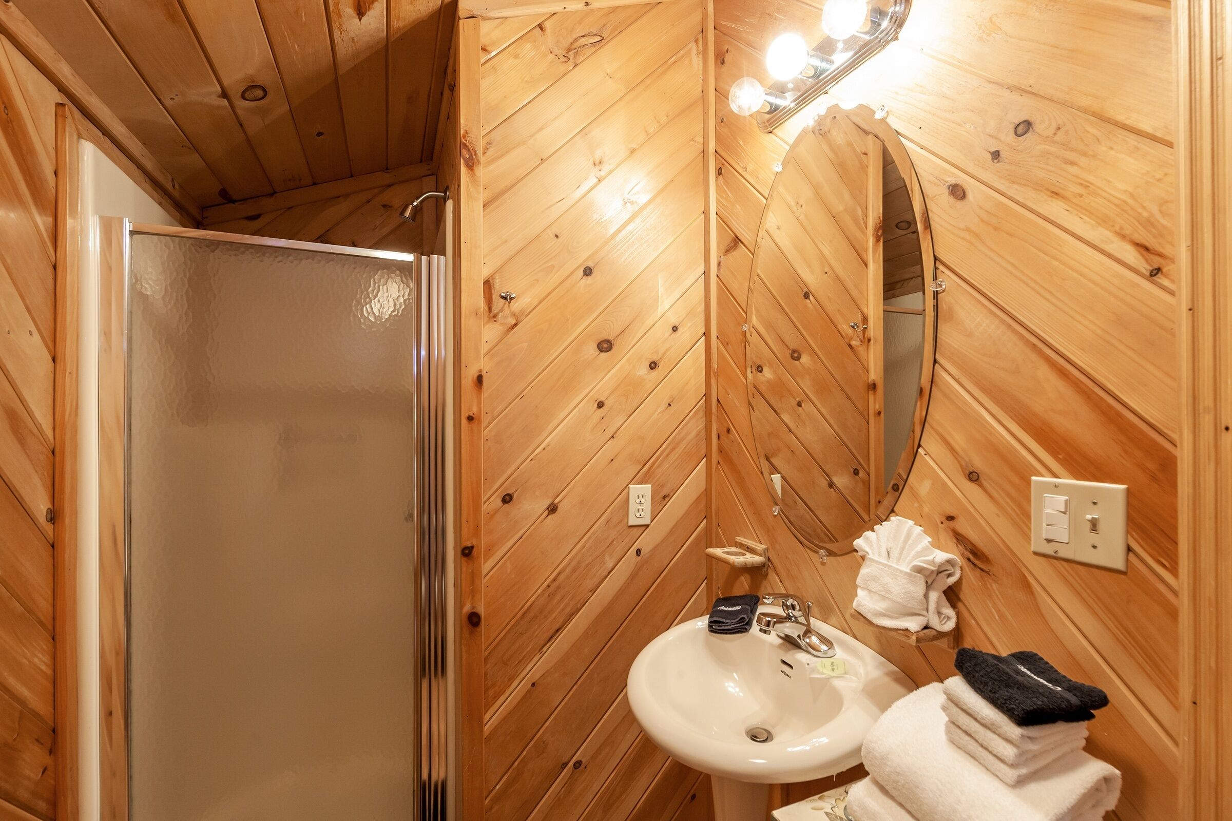 Un baño revestido de paneles de madera con cabina de ducha de vidrio, un lavamanos y un espejo.