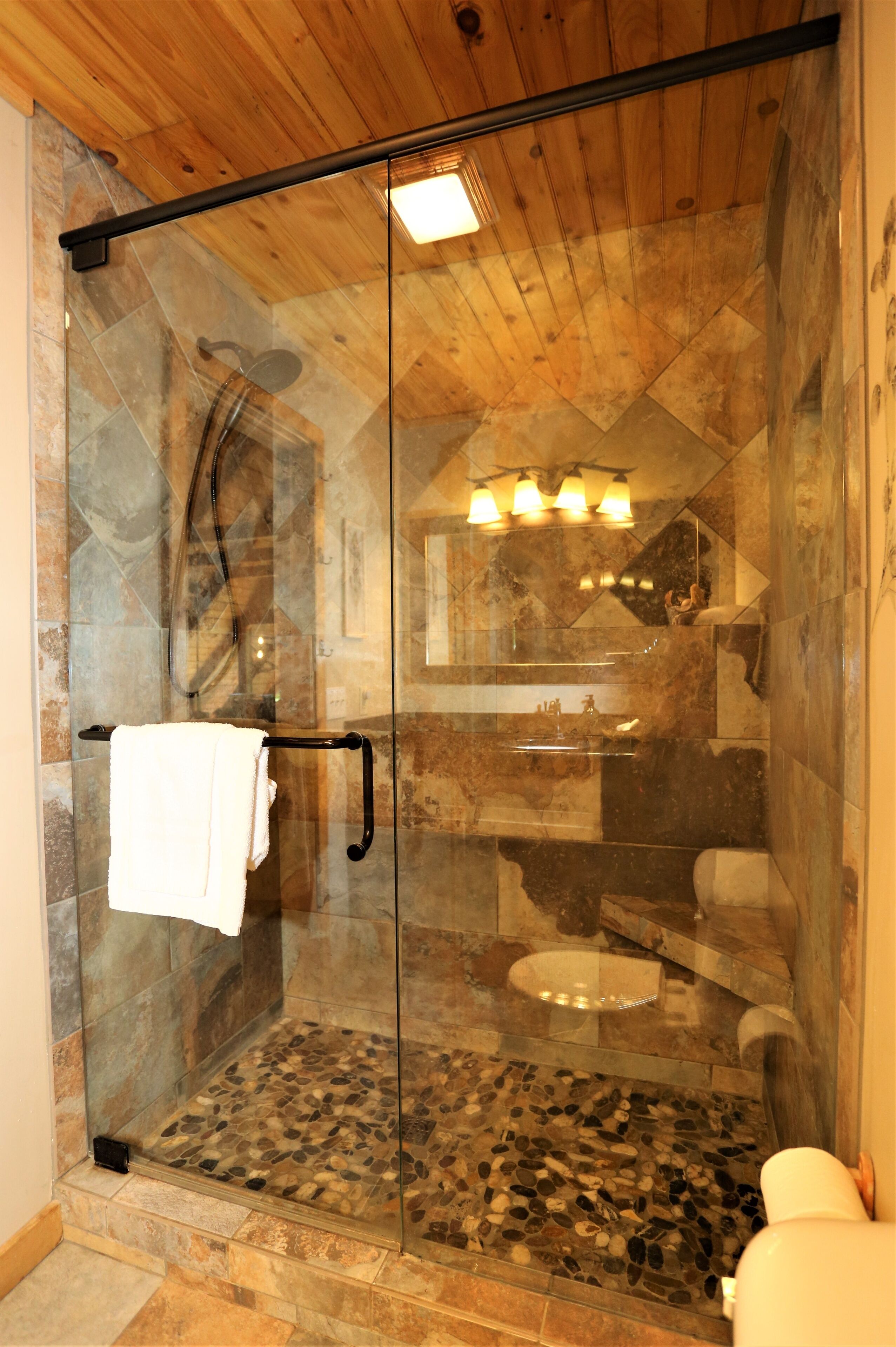 A glass-enclosed shower with a stone floor, a wooden ceiling, and a visible bathroom sink and toilet.