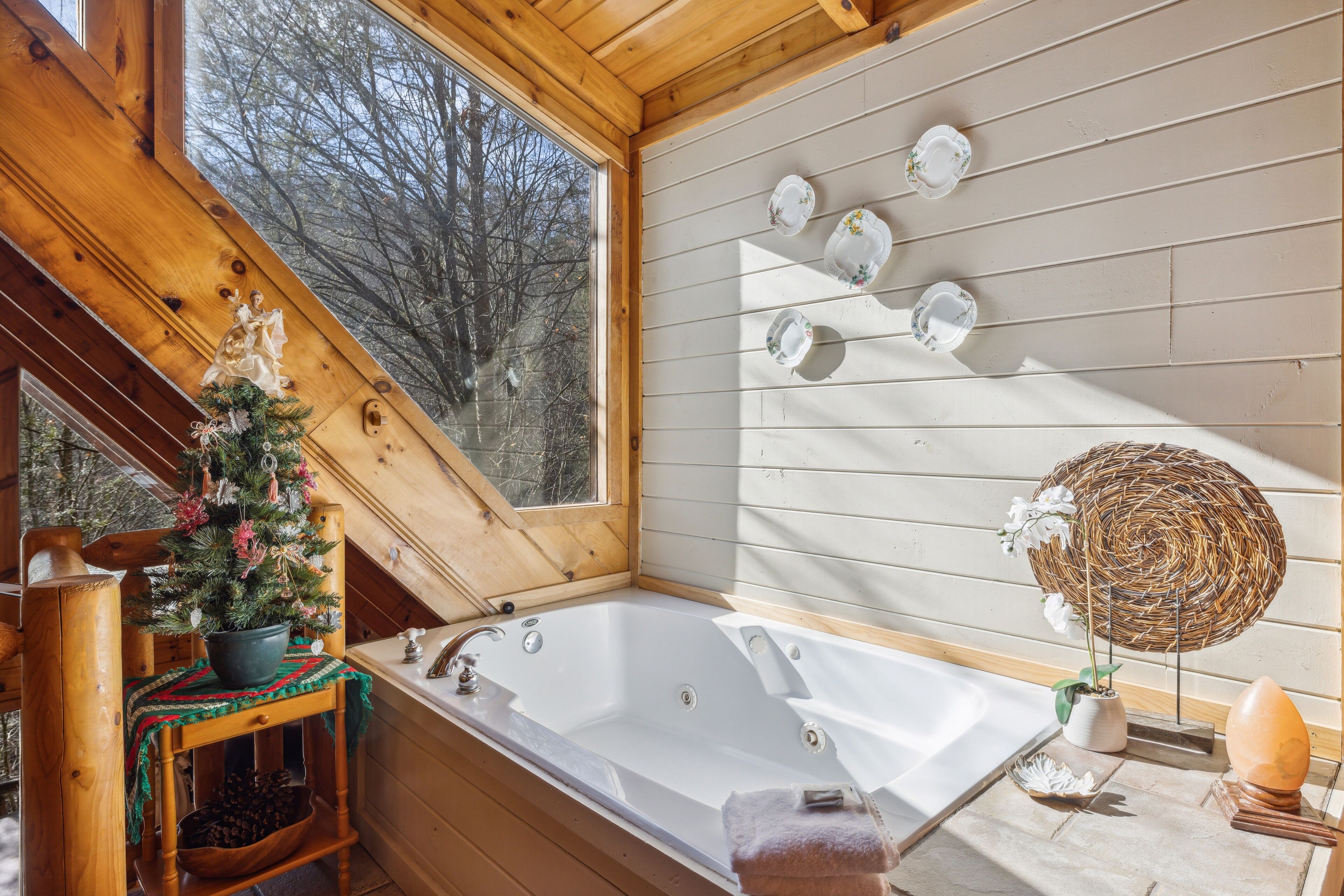 A wooden cabin interior with a large bathtub, a Christmas tree, and a view of trees through a window.