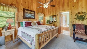 Quarto de cabana de madeira com cama, cadeira, mesa de cabeceira, abajur, ventoinha no teto e porta que dá para outro cômodo.