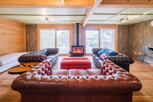 Un salón acogedor con sofás de cuero, una chimenea central y ventanas amplias que ofrecen vistas al exterior.
