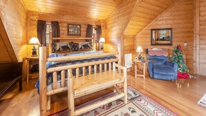 A cozy log cabin bedroom with a wooden bed, two nightstands, a small table, a blue armchair, a Christmas tree, and a mirror on the wall. - Something Extra 2 Bedroom Cabin by RedAwning (Sevierville)