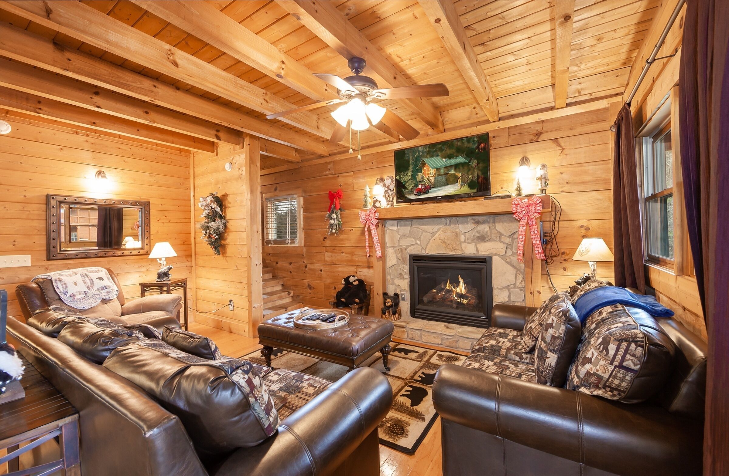 A cozy log cabin interior with a fireplace, leather sofas, a television, and a ceiling fan.