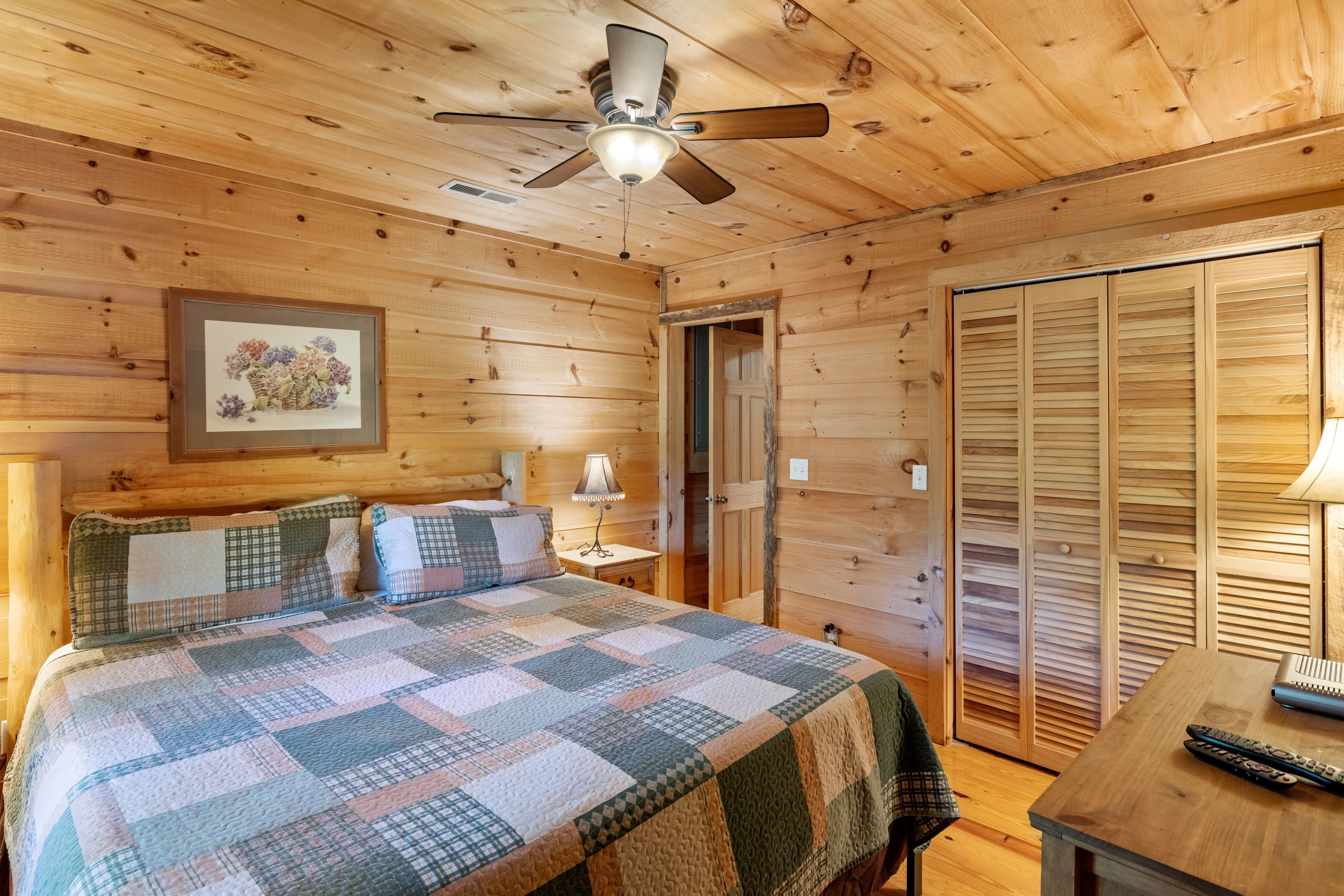 Une chambre dans une cabane en bois, avec un lit, une table de chevet, un ventilateur de plafond et une armoire en bois.