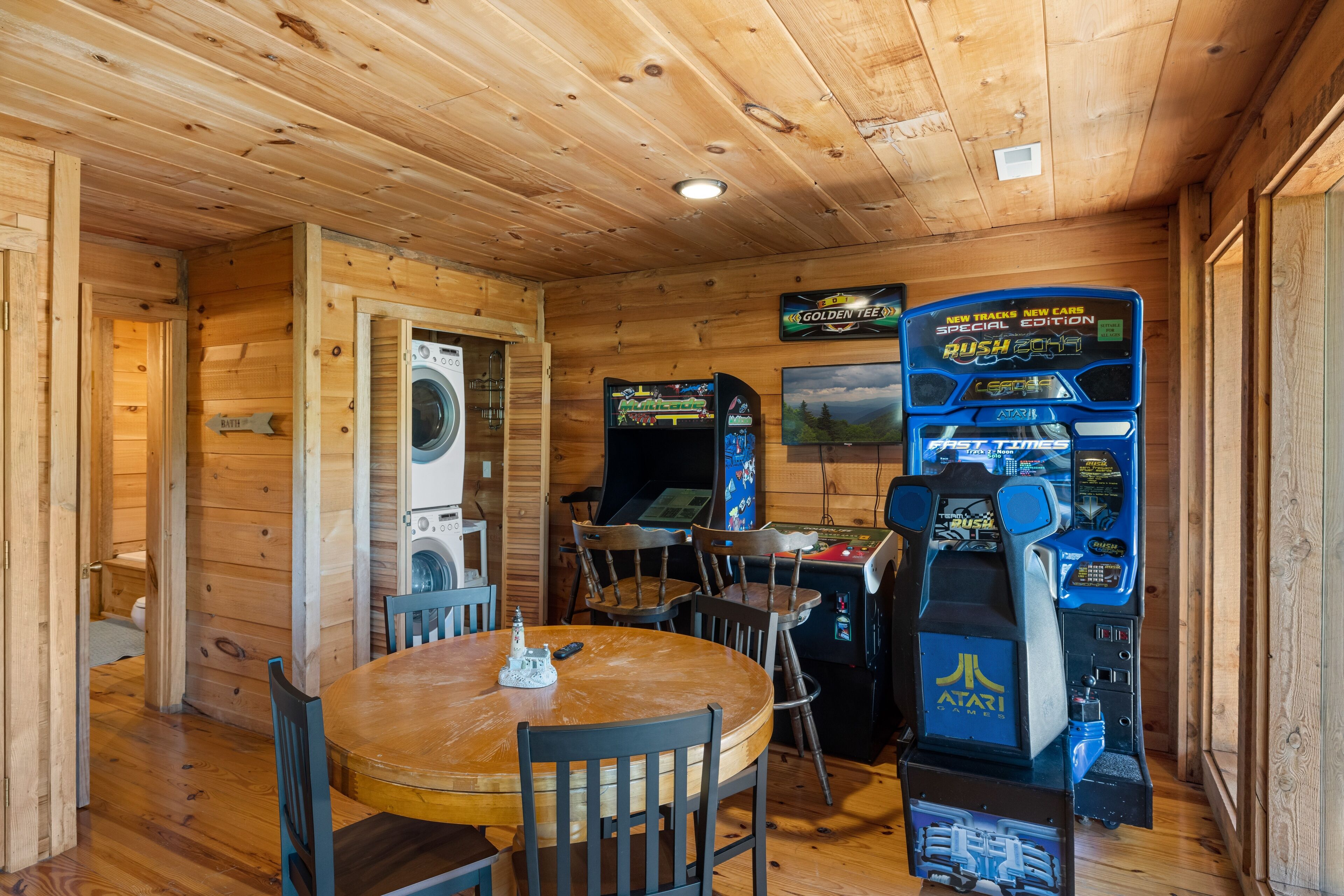 A wooden interior with a round table, chairs, a washing machine, and arcade games.