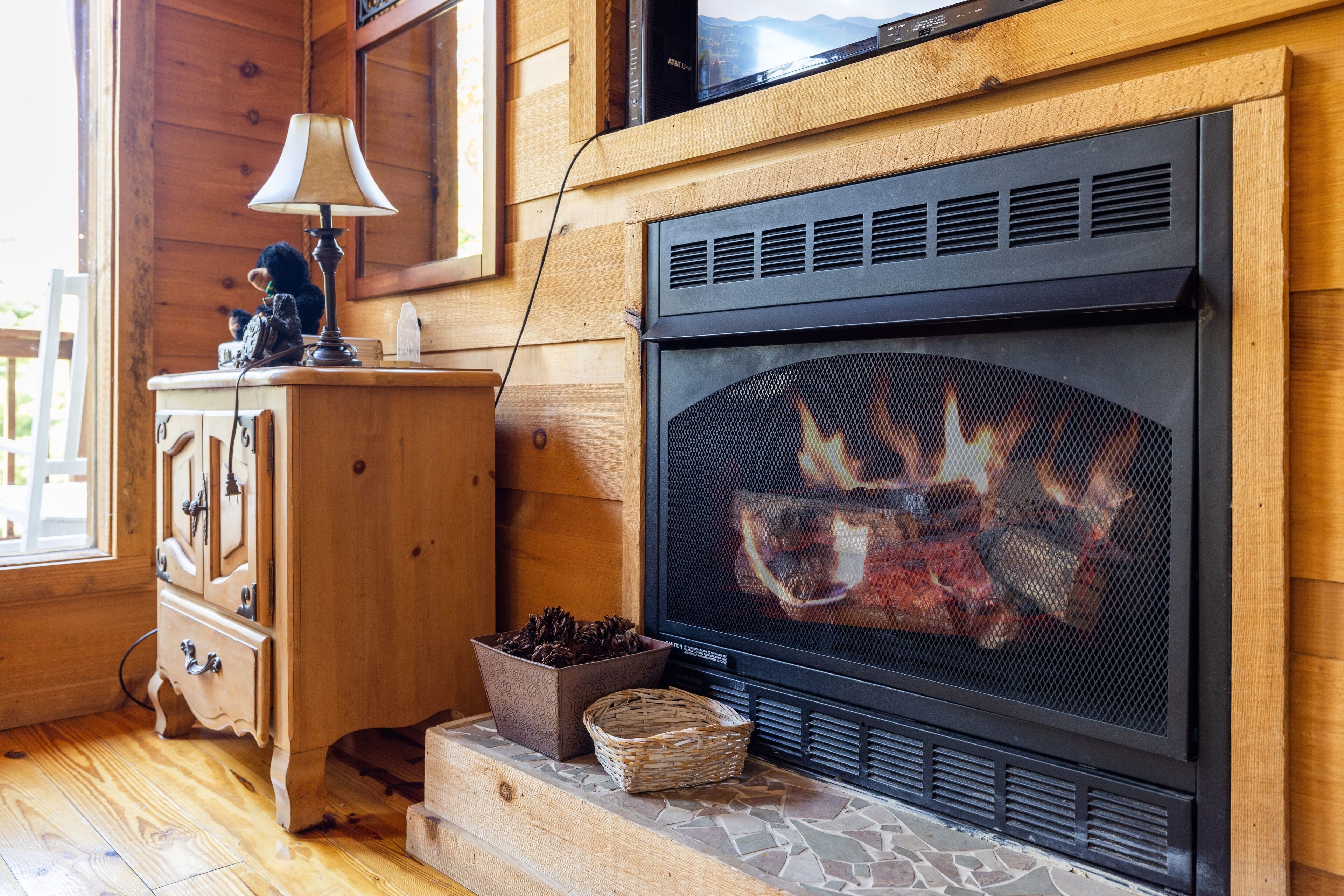 Una chimenea con protección, una lámpara y un aparador de madera con cajones.