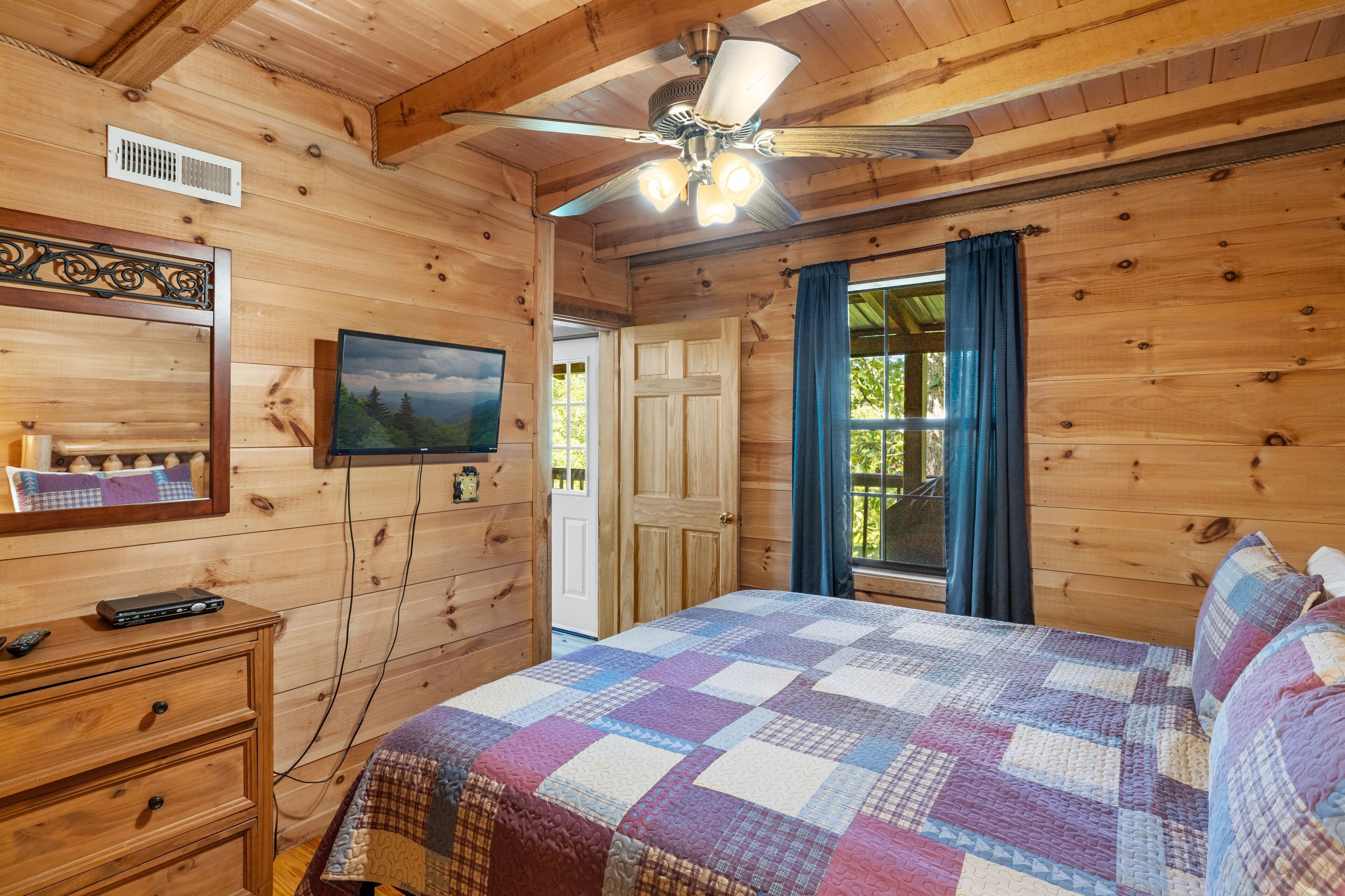 A wooden cabin bedroom with a bed, dresser, ceiling fan, and a TV on the wall.