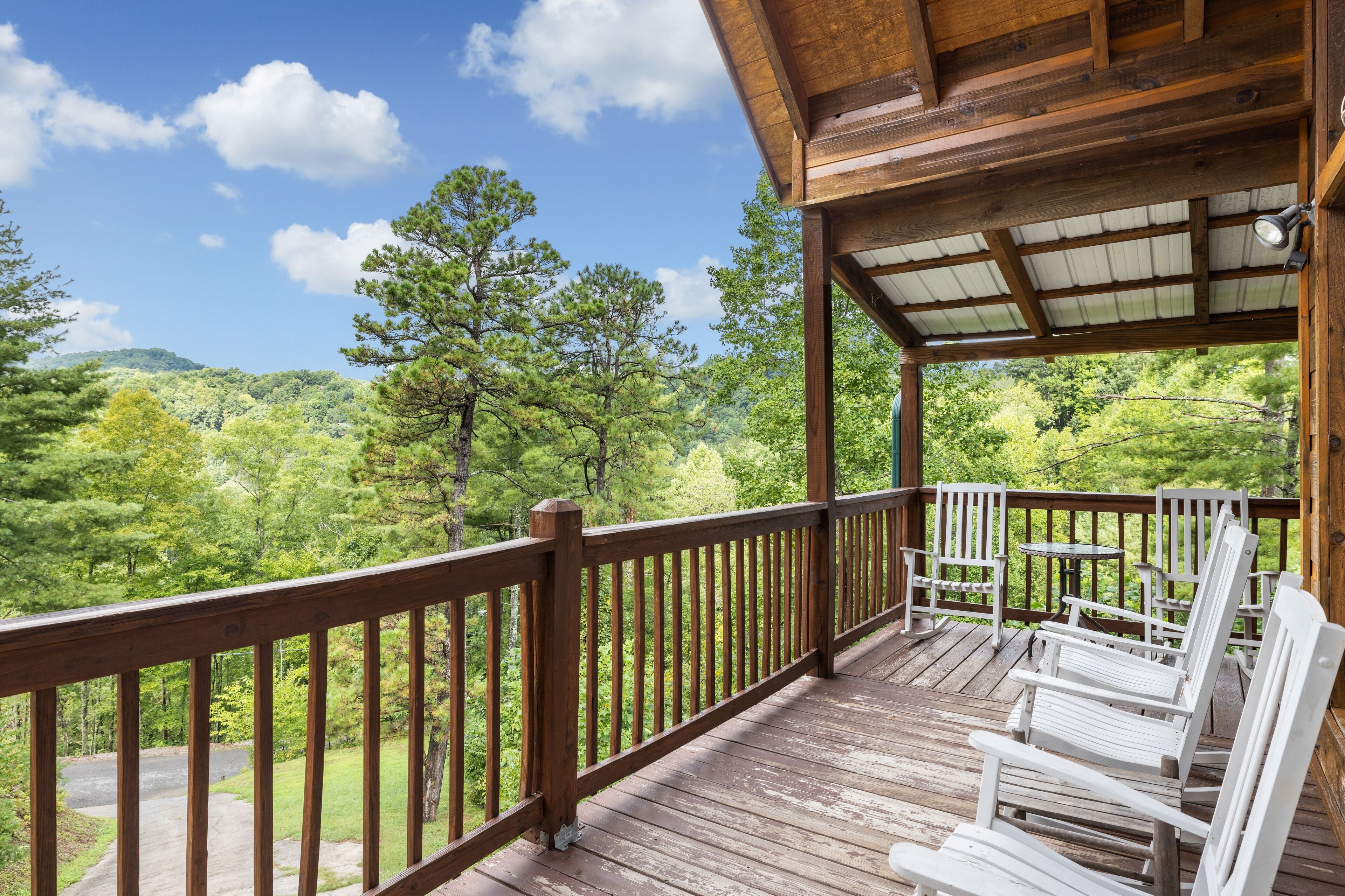 A wooden deck with white chairs and a table, overlooking a forested landscape.