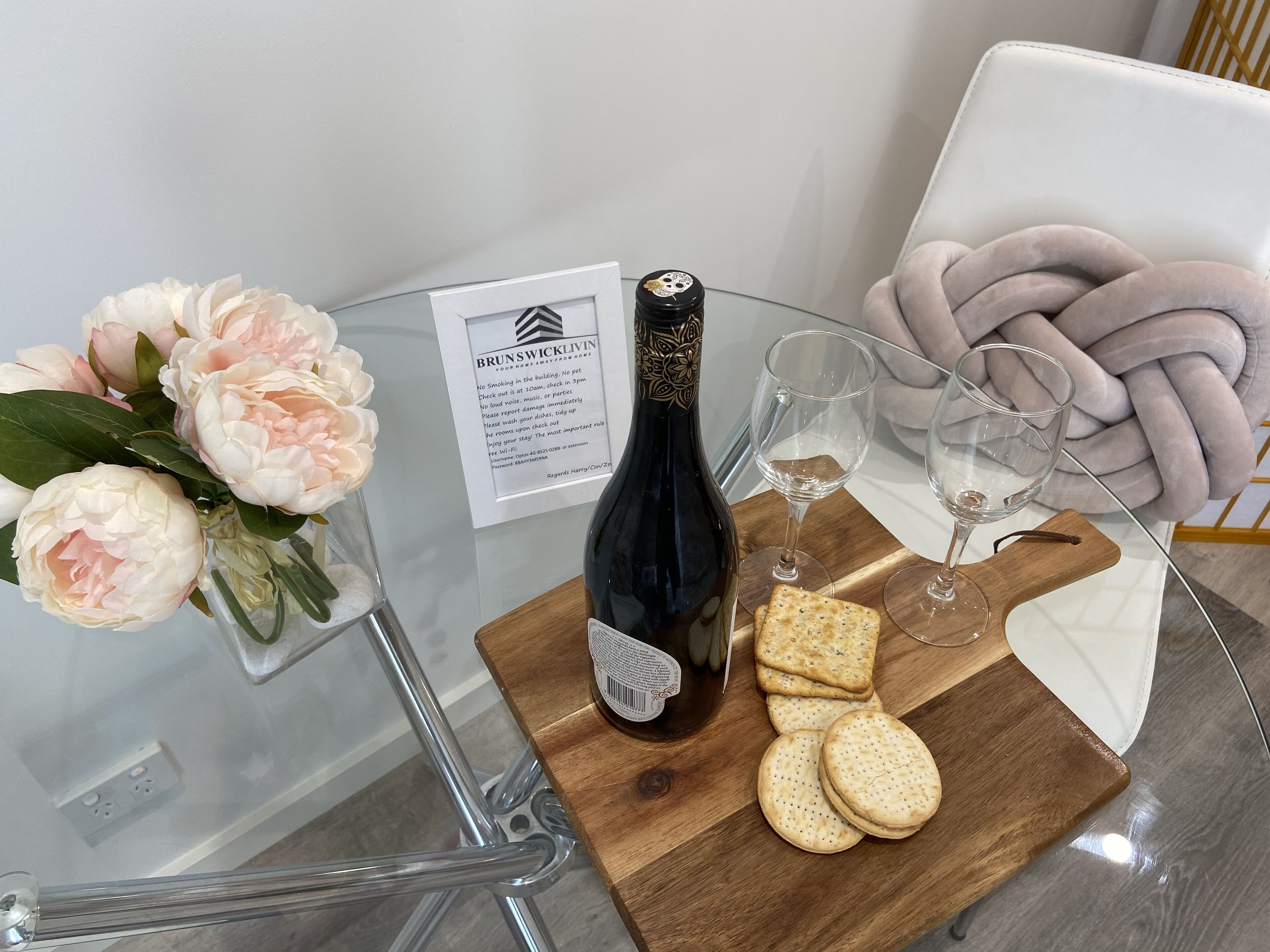A glass table with a bottle of wine, glasses, crackers, and a bouquet of flowers.