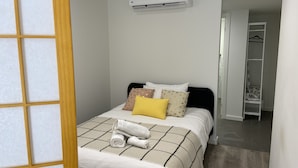Un letto rifatto con cura, con una coperta a quadri, un asciugamano arrotolato e diversi cuscini, incluso uno giallo. Un condizionatore d'aria è montato sulla parete sopra il letto.