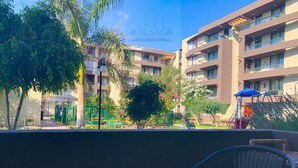 A balcony with wicker chairs, a potted plant, and a view of a multi-story building with balconies and greenery. - Apartment Alma Surire pool view (Arica)