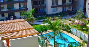 A rooftop pool area with a wooden pergola, surrounded by lush greenery and palm trees.
