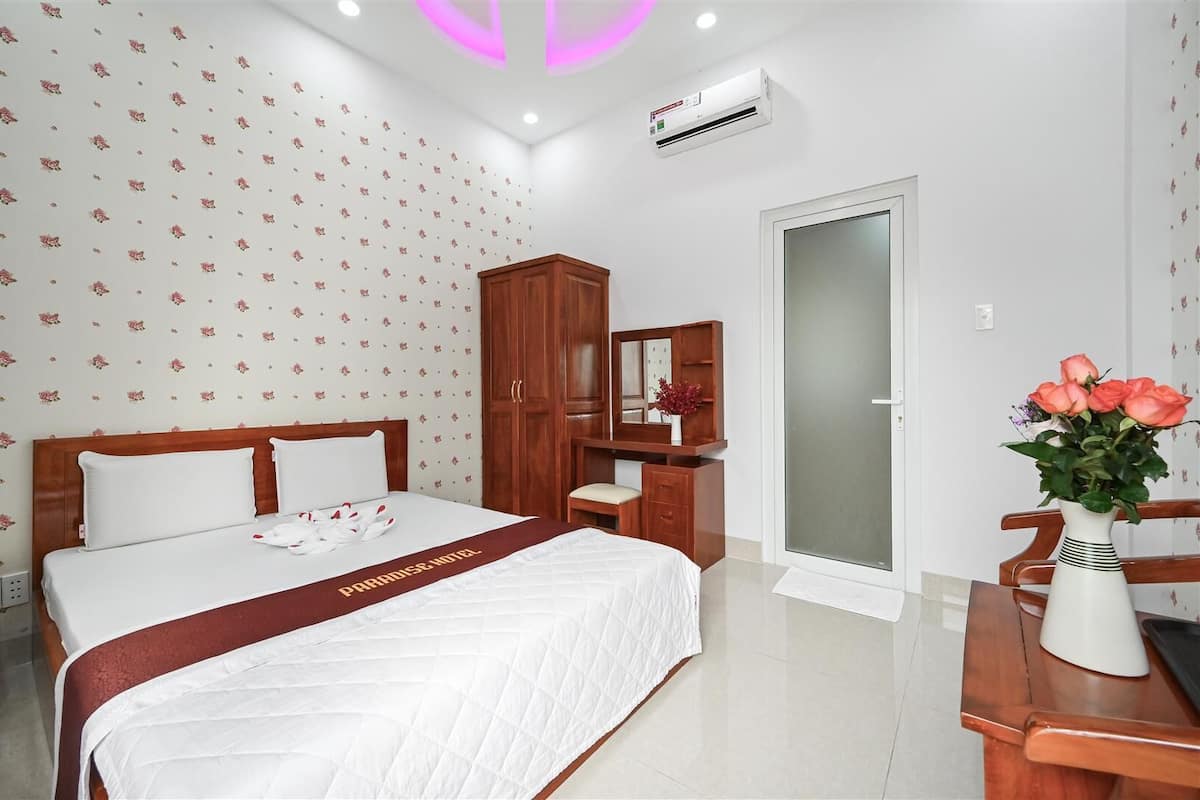 A hotel room with a bed, a wooden wardrobe, a vanity with a mirror, a desk, and a vase with flowers.