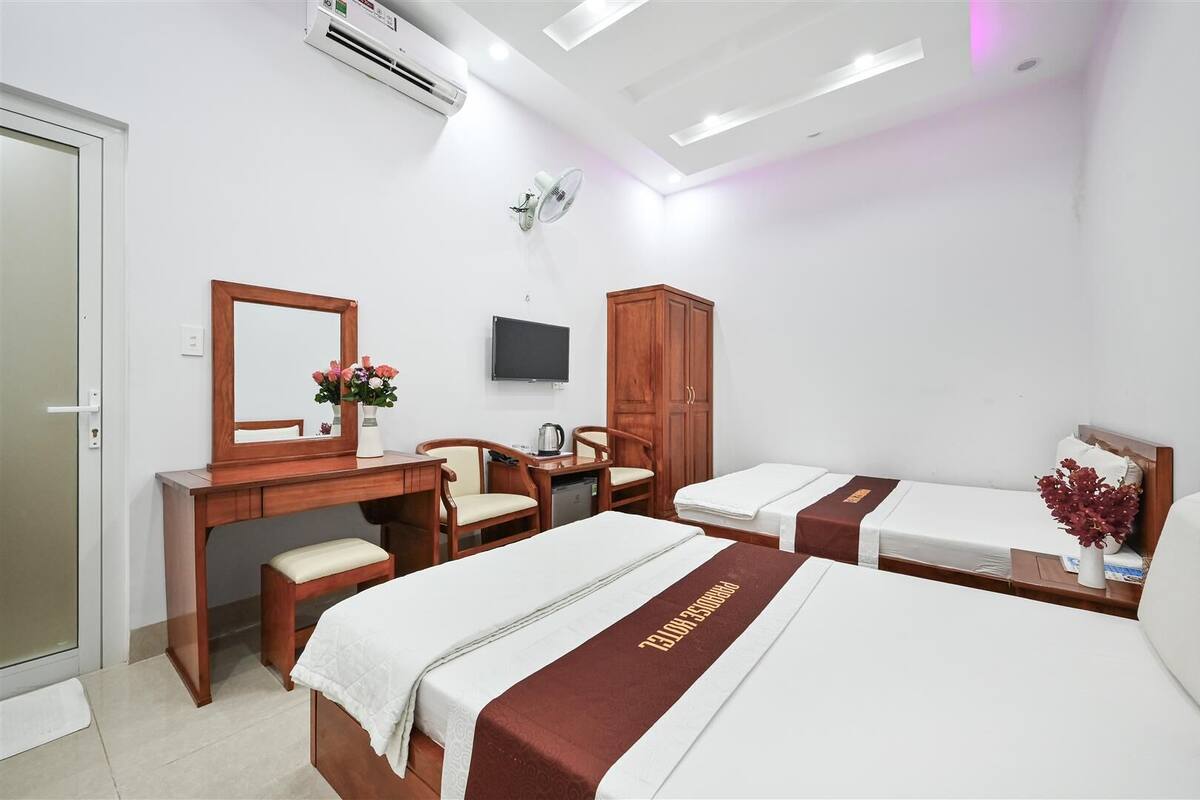 A hotel room with two single beds, a wooden desk with a chair, a wardrobe, a television, and a small table with a vase of flowers.