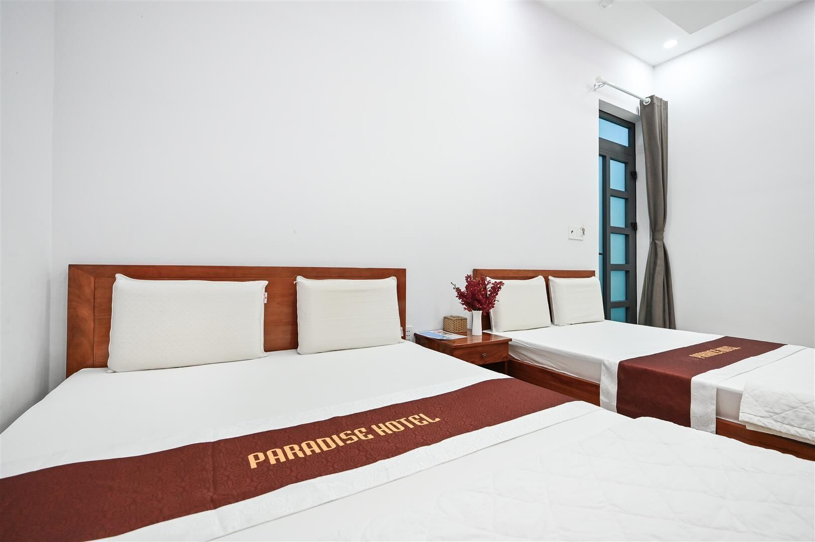 A hotel room with two beds, each with a wooden headboard and white bedding. A red and gold bed runner with the text "PARADISE HOTEL" is visible.